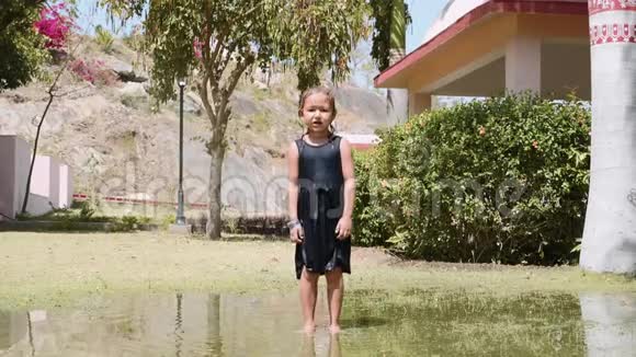 穿着衣服的快乐湿赤脚女孩在公园的水坑里跳视频的预览图