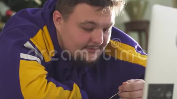 大胡子的年轻人坐在椅子上看着笔记本电脑用勺子吃果酱视频的预览图