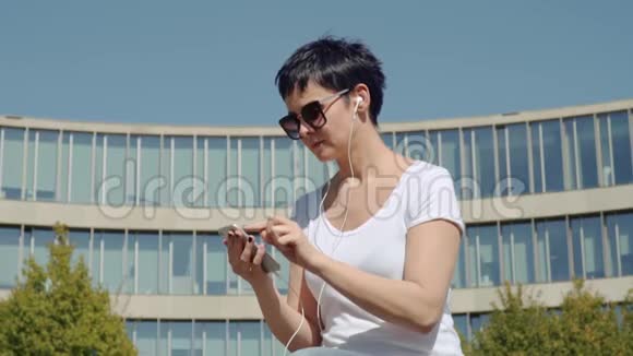 商务女性穿着白色T恤坐在办公楼前听音乐视频的预览图