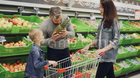 父亲母亲和孩子在杂货店里选择菠萝摸着它闻着它竖起大拇指货架视频的预览图