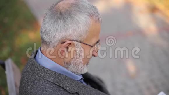 英俊的白发老人长着美丽的胡须坐在长凳上用平板电脑在网上滚动视频的预览图