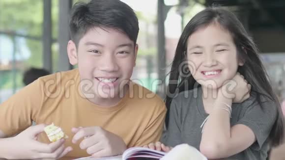 亚洲男孩和女孩用卡通书放松可爱的亚洲孩子坐在咖啡馆里享受读书微笑着面对视频的预览图