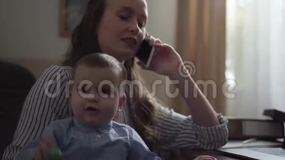 忙碌的年轻母亲在家工作的肖像带着婴儿在她的大腿上那个女人在儿子的时候用手机说话视频的预览图