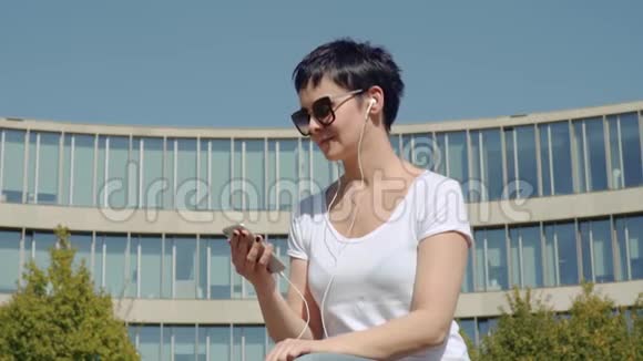 商务女性穿着白色T恤坐在办公楼前听音乐视频的预览图