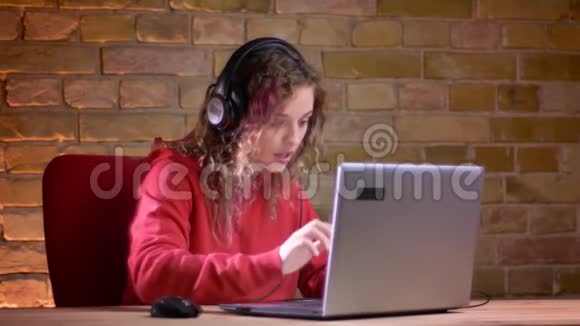 穿红色连帽衫的年轻女性博客写手的肖像很高兴在布里克肯墙背景上使用笔记本电脑交谈视频的预览图