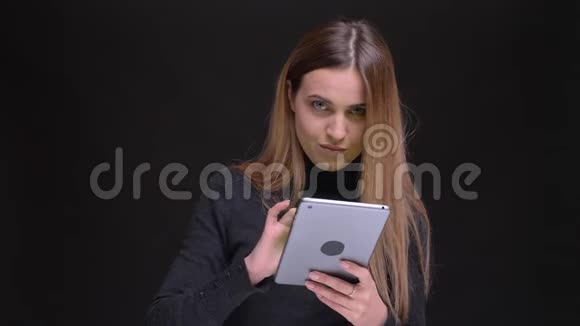 年轻的长发金发金发女孩的肖像微笑着将绿色的平板电脑屏幕显示在黑色背景下的相机中视频的预览图
