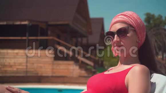 一个漂亮的怀孕女孩穿着红色的一件泳衣和眼镜躺在游泳池旁的休息室里视频的预览图