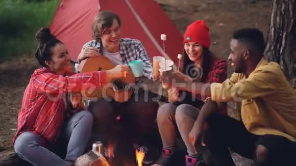快乐的年轻人朋友们正在森林里围着火堆手里拿着温暖的棉花糖手里拿着酒杯和饮料视频的预览图
