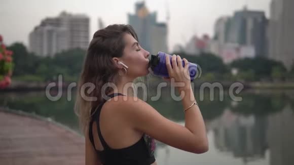 年轻的黑发女人在公园里跑步后在空中脚听音乐在蓝色健身杯里喝健身饮料视频的预览图