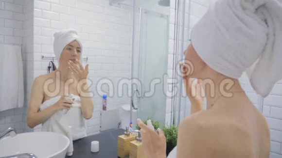 年轻的女人拿着毛巾脸上涂着奶油在家里的浴室里看着镜子美容生活方式护肤理念视频的预览图