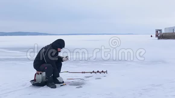 穿着深色衣服的老渔夫在冰冻的河上钓鱼喝热茶或咖啡视频的预览图