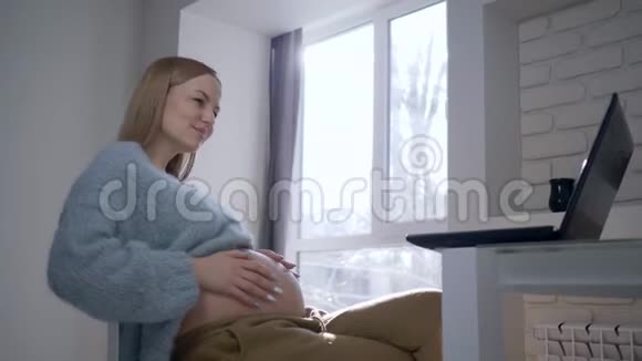 怀孕的女孩带着笔记本电脑坐在桌子旁背光在家里抚摸她赤裸的肚子视频的预览图