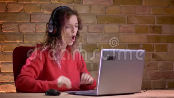 穿着红色连帽衫的年轻女性博客写手在布里克肯墙上愉快地拍手和用笔记本电脑聊天的画像视频的预览图