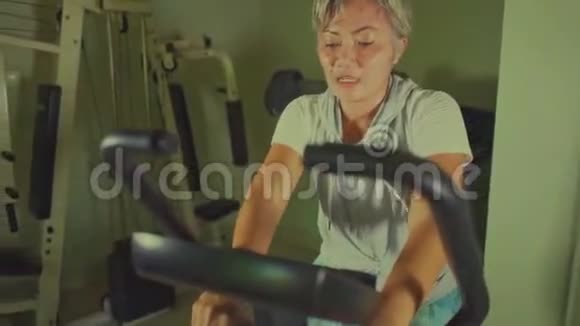 一辆万向节静态自行车在中年迷人和出汗的印度尼西亚妇女身上拍摄她的白发训练在健身房出汗视频的预览图