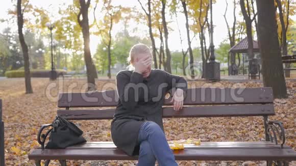 女人哭着捂着脸手掌坐在秋天公园的长凳上视频的预览图