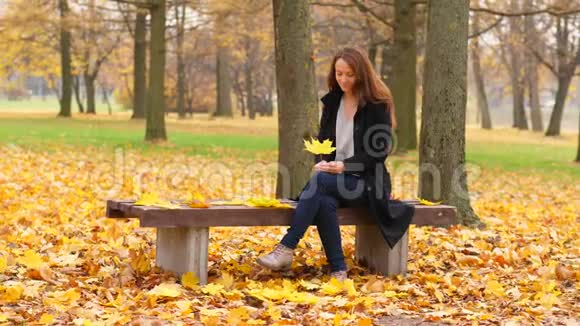 女人坐在秋天的公园里与落叶嬉戏视频的预览图