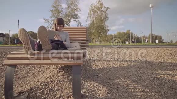 在凉爽的一天带着平板电脑的女孩坐在鹅卵石海滩上的木制甲板椅上视频的预览图
