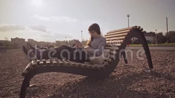 在凉爽的一天带平板电脑的自由职业者坐在鹅卵石海滩上的木制甲板椅上视频的预览图