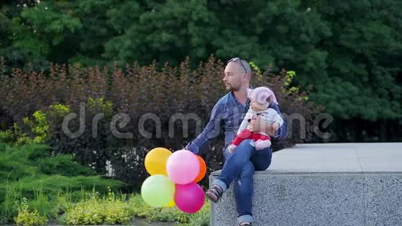 爸爸手里拿着五颜六色的气球可爱的女儿坐在画城公园的水泥墙上视频的预览图