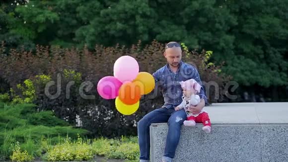 爸爸手里拿着五颜六色的气球可爱的女儿坐在画城公园的水泥墙上视频的预览图