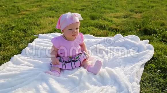 可爱漂亮的女婴穿着粉红色的衣服坐在铺在城市公园绿草上的白色毯子上视频的预览图