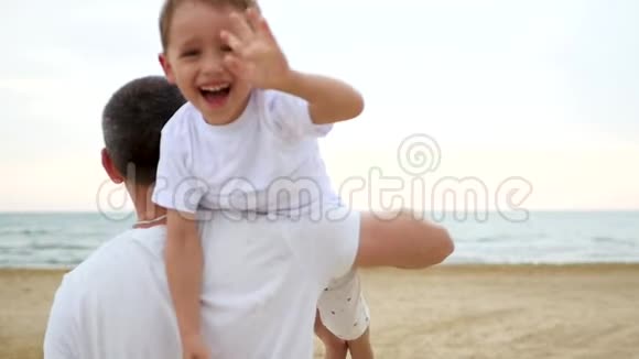 父子俩在沙滩上玩耍那个人肩上扛着一个快乐的笑着的男孩挥舞着手臂视频的预览图