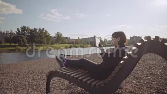 在凉爽的一天带着平板电脑的女孩坐在鹅卵石海滩上的木制甲板椅上视频的预览图