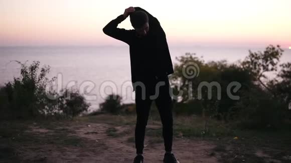 身穿黑色运动服的黑发青年男子正在热身站在草地上伸展双臂美丽的景色视频的预览图