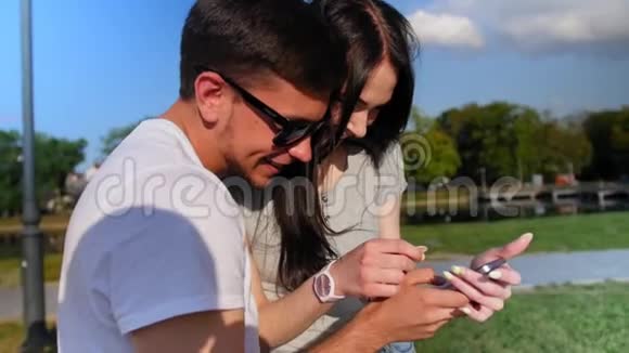 站在公园里的年轻漂亮的夫妇使用现代手机男人在他的智能手机上显示一些东西女孩视频的预览图