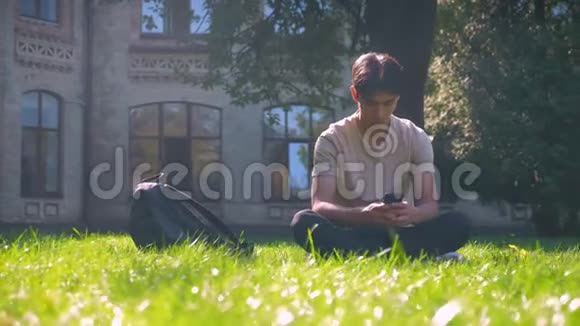 英俊的亚洲男人坐在阳光下的草地上滚动着他的手机专注的城市视野研究着气氛视频的预览图