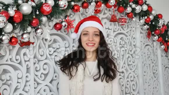 圣诞装饰的摄影棚里戴着红色圣诞帽的漂亮微笑的年轻女子看镜头的画像视频的预览图
