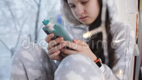 穿着温暖睡衣的少女穿着冬天的窗户在智能手机上看书视频的预览图