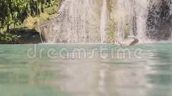 年轻女子从飞溅的瀑布中在蓝色的湖水中游泳快乐的女人在山湖和水中游泳视频的预览图