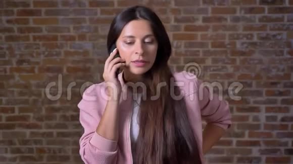 一位深褐色头发的白种人女性集中在她的电话交谈中站在砖墙旁边交谈视频的预览图