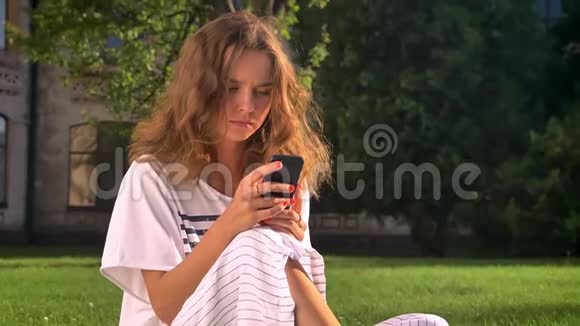 年轻的白种人黑发女人坐在草地上的公园里用智能手机滚动打字背景是大学视频的预览图