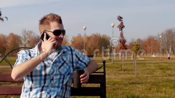 年轻时尚时髦的戴着黑色眼镜的男人坐在长凳上喝咖啡在电话特写和复制上交谈视频的预览图