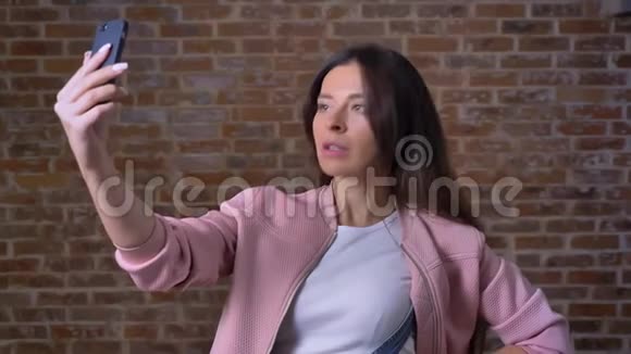 穿着粉红色夹克的白种人漂亮的女人正拿着她的智能手机自拍一边站在红色的地方一边显示着亲吻标志视频的预览图