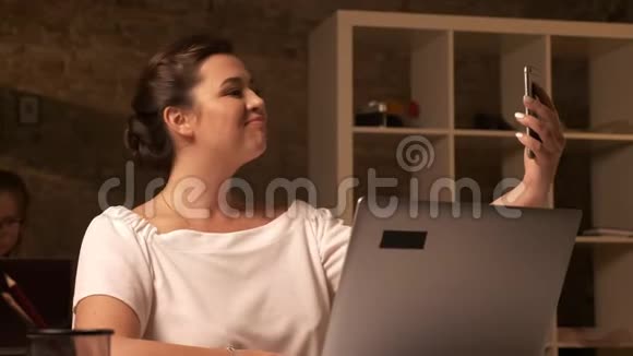 可爱的高加索女商人正坐在棕色工作室笔记本电脑附近的另一个女人旁边自拍视频的预览图
