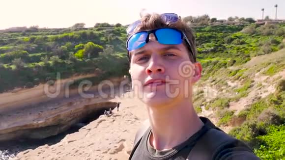 年轻的蓝眼男带着两副太阳镜在大自然海湾绿岩的背景下自拍视频的预览图
