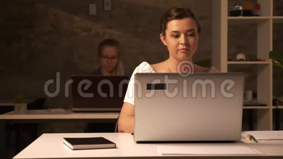 两个工作的白种人妇女坐在棕色的办公室里在室内的白色办公桌上使用笔记本电脑视频的预览图