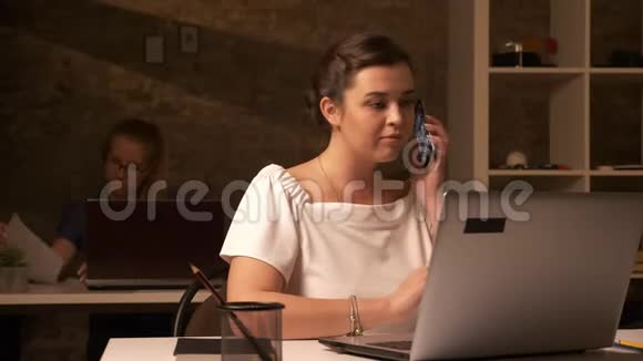 美丽的白种人女孩坐在她的同事旁边的白色办公桌旁用电话交谈视频的预览图