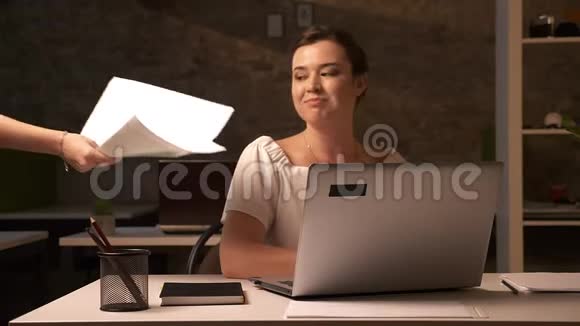 快乐的白种人女人坐在靠近她的笔记本电脑的办公室里一边写着工作文件一边放松视频的预览图