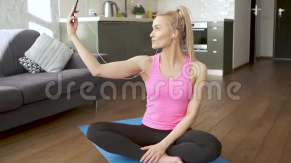 年轻迷人的运动女性坐在瑜伽垫上用手机自拍视频的预览图