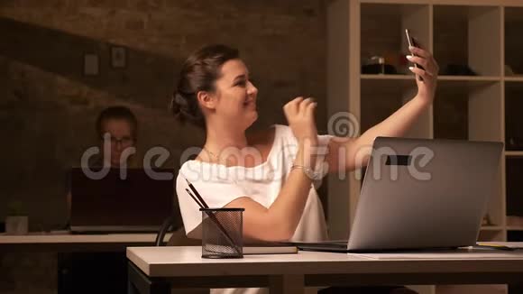 伟大的白种人女性正在智能手机上自拍而她正和同事坐在一起办公桌分开视频的预览图