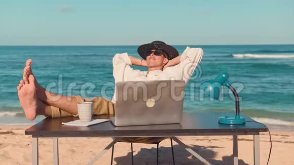 一个年轻人拿着笔记本电脑坐在海滩上休息着把腿放在桌子上视频的预览图