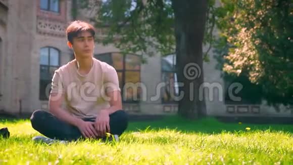 漂亮的亚洲男孩在坐在草地上时用耳机听音乐而建筑物在后面是孤立的视频的预览图