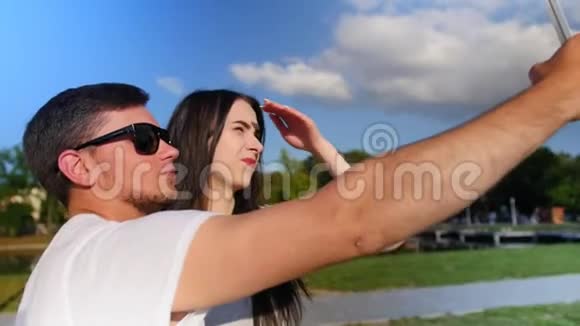 中镜头的快乐年轻夫妇自拍与智能手机同时站在公园附近的池塘近距离射击视频的预览图