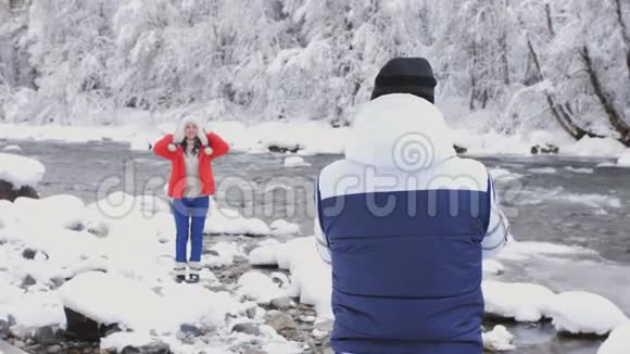 美丽的女孩穿着冬天的夹克和帽子为摄影师摆姿势冰肆虐的冬季河流雪景视频的预览图