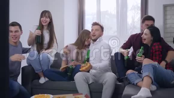 快乐的朋友坐在电视机前的沙发上吃披萨在家庭聚会上用啤酒瓶做烤面包视频的预览图