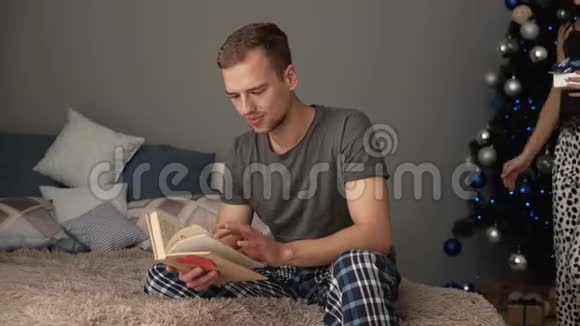 英俊的白种人穿着家居服坐在床上看书一个年轻人过圣诞节视频的预览图